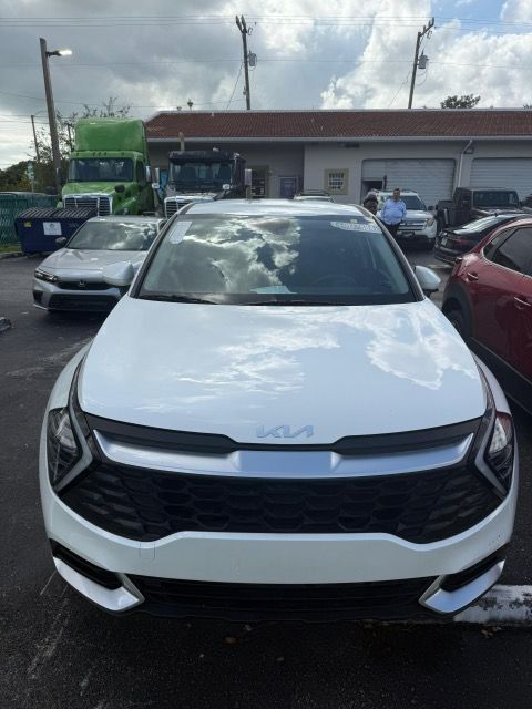 White Kia SUV parked outdoors with other vehicles and a green truck in the background.