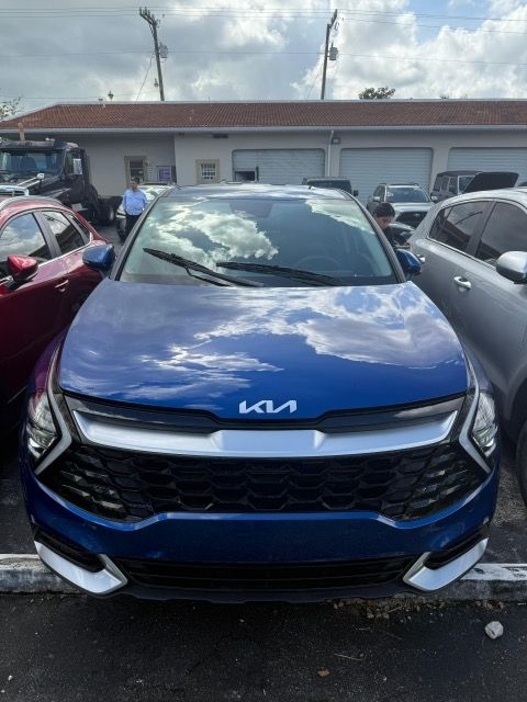 Blue Kia SUV parked in front of a building with white garage doors; cloudy sky overhead.