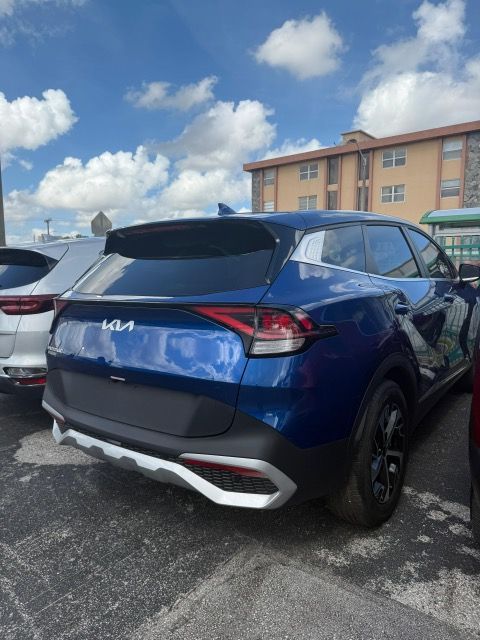 Blue Kia Sportage SUV parked, viewed from the rear, with a building and blue sky in the background.