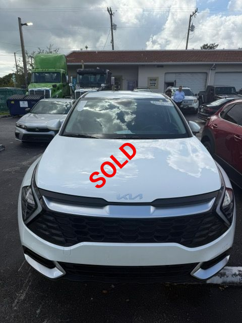 White Kia SUV parked outdoors with other vehicles and a green truck in the background.