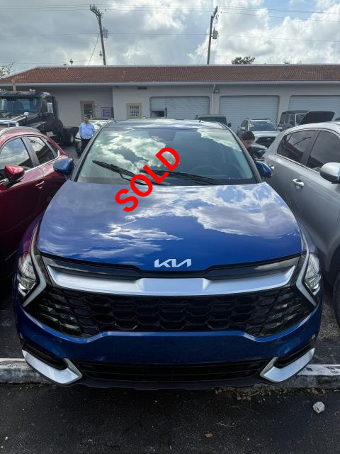 Blue Kia SUV parked in front of a building with white garage doors; cloudy sky overhead.