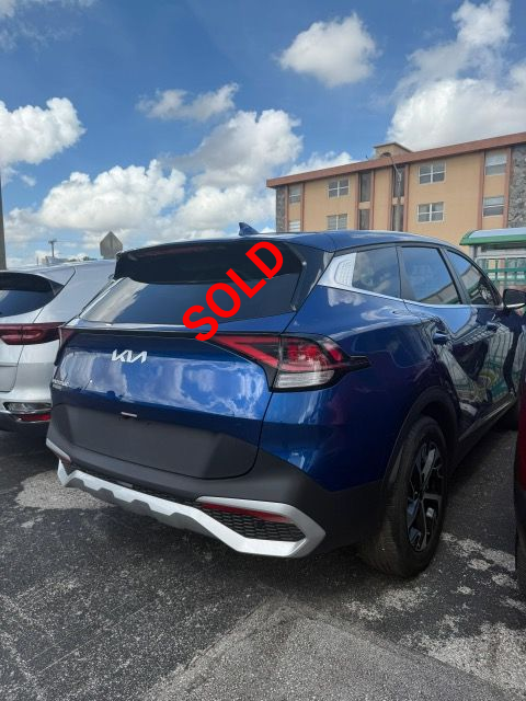 Blue Kia Sportage SUV parked, viewed from the rear, with a building and blue sky in the background.