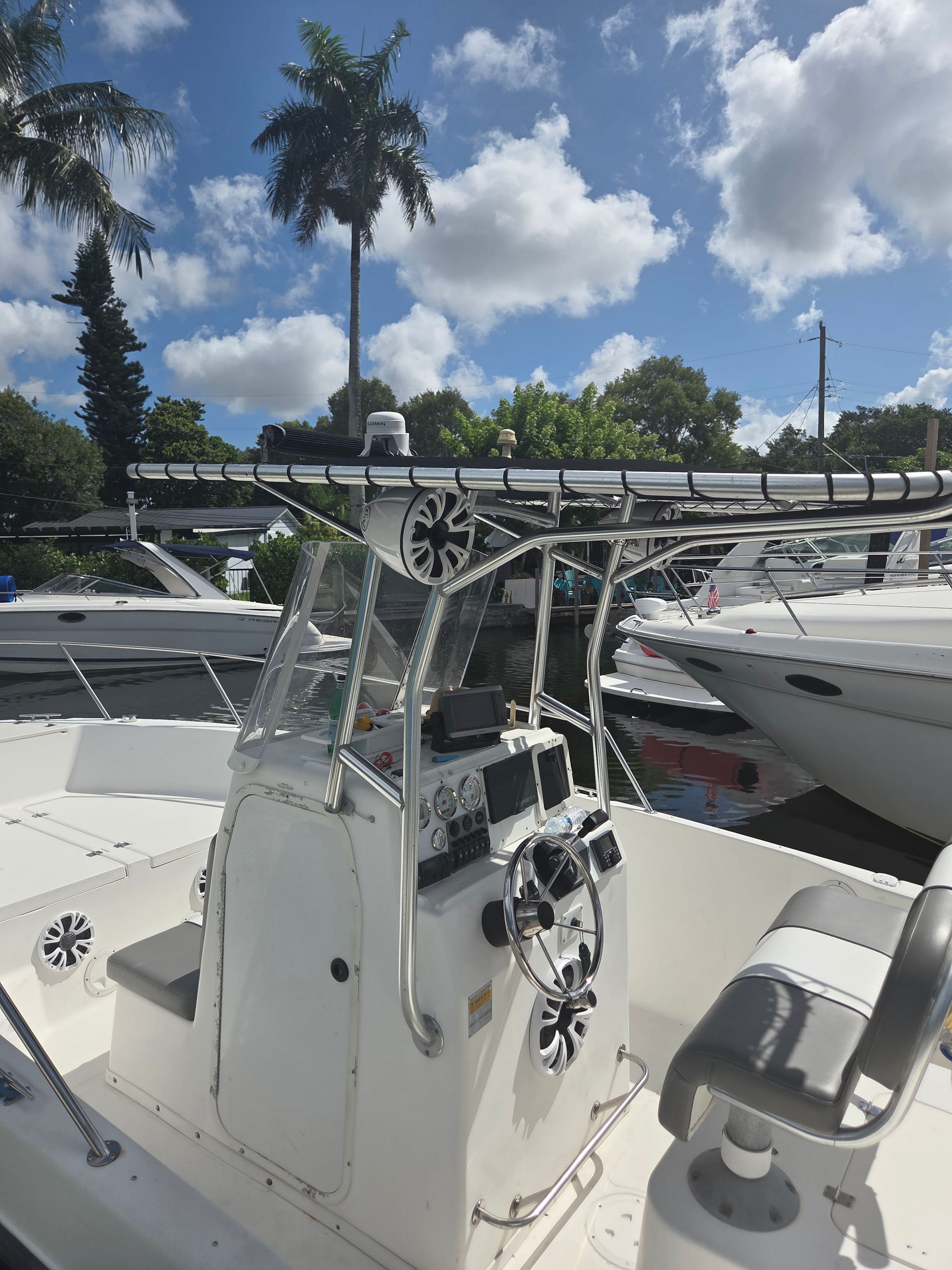 White boat's helm with clear windshield, steering wheel, and gauges. Palm tree, bridge, and sky in the background.