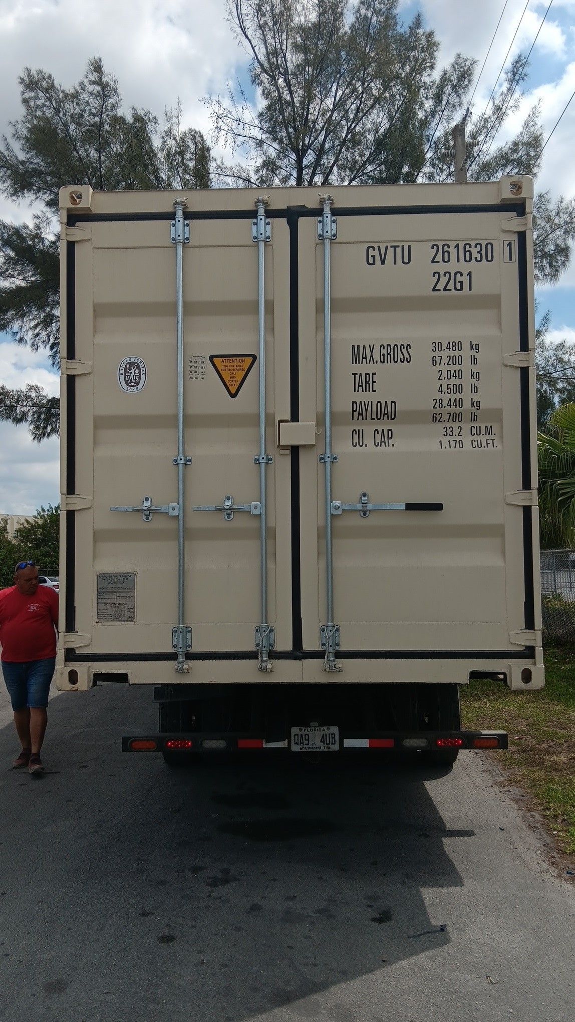 Tan cargo container on a truck, man in red shirt stands nearby on asphalt road.