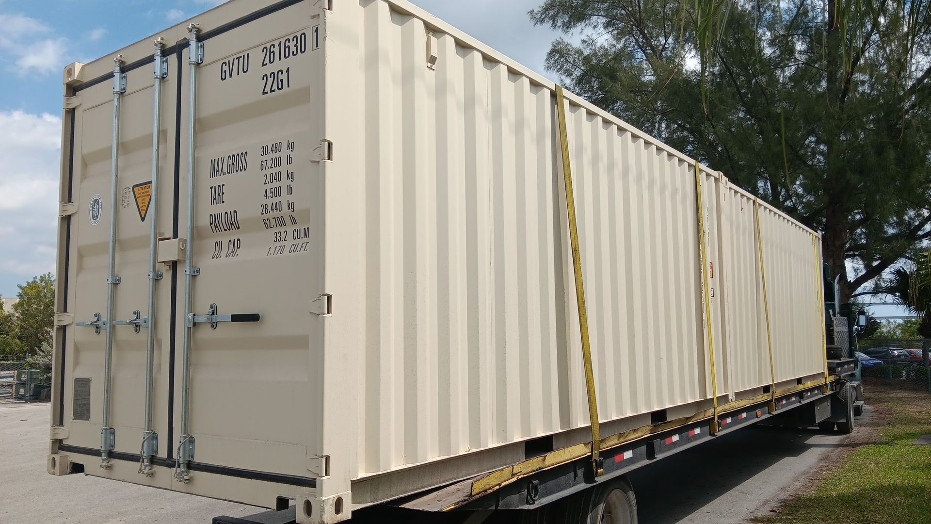 Beige shipping container on a flatbed truck, secured by yellow straps, against a blue sky.