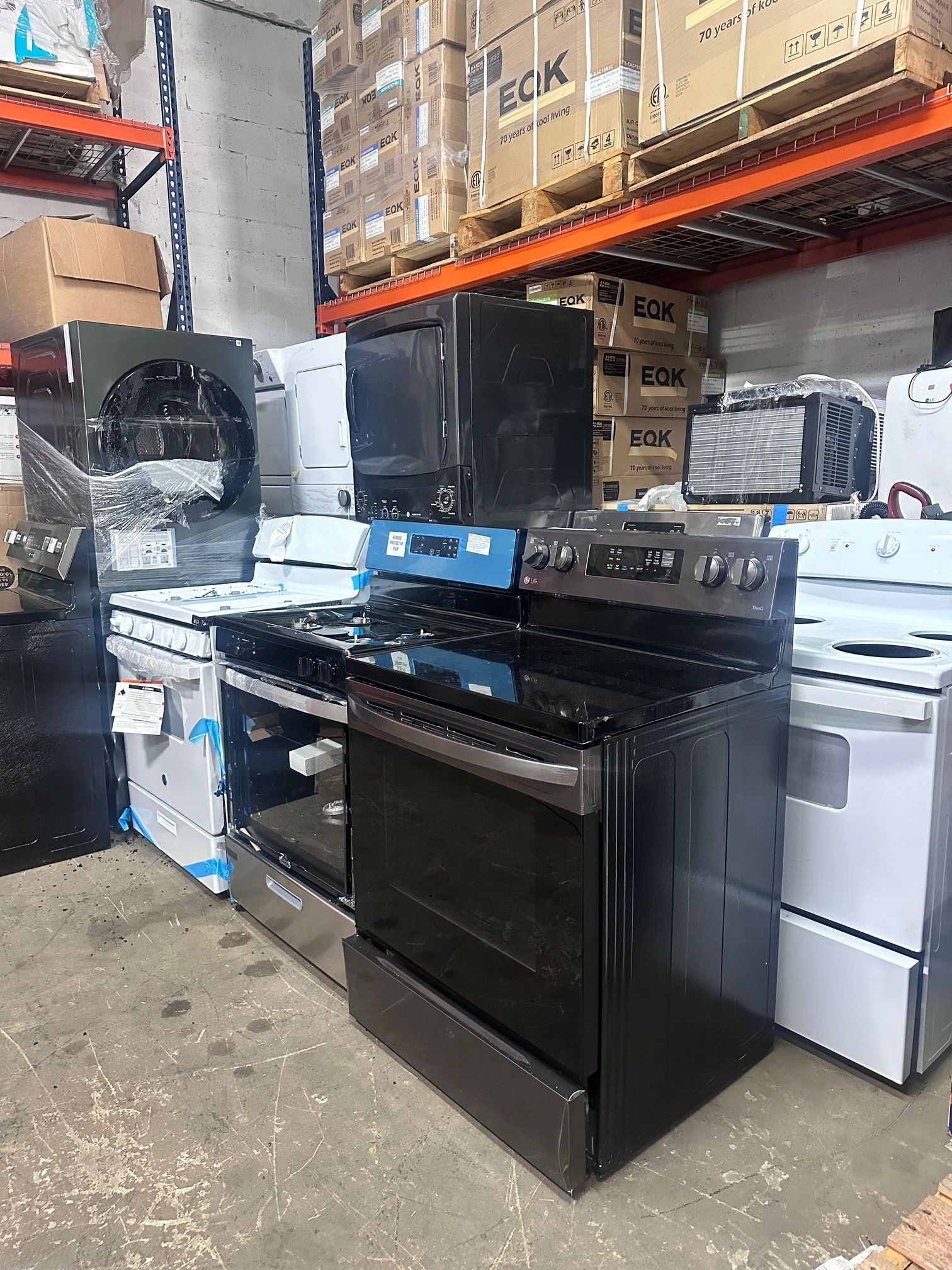 Rows of kitchen appliances in a warehouse, including ovens and a microwave, stacked near pallets of boxed goods.