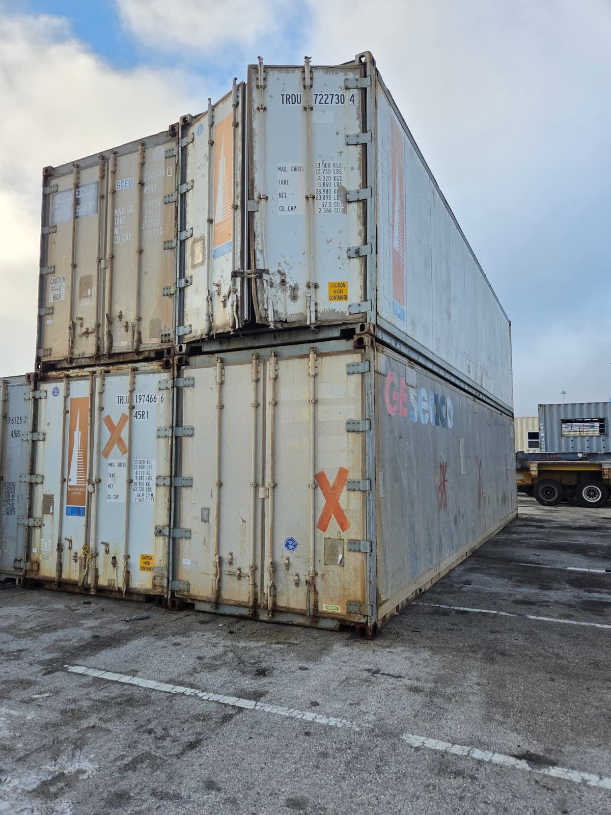 Three stacked shipping containers, weathered and gray, on an asphalt lot under a cloudy sky.