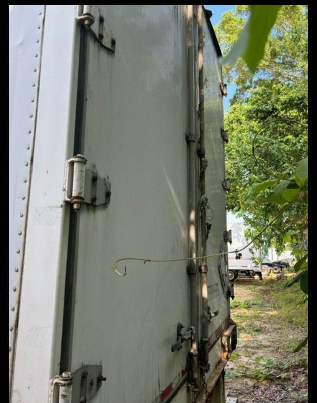 Side view of a white semi-trailer. Gray metal latches and hinges. Green foliage surrounds it in a natural setting.