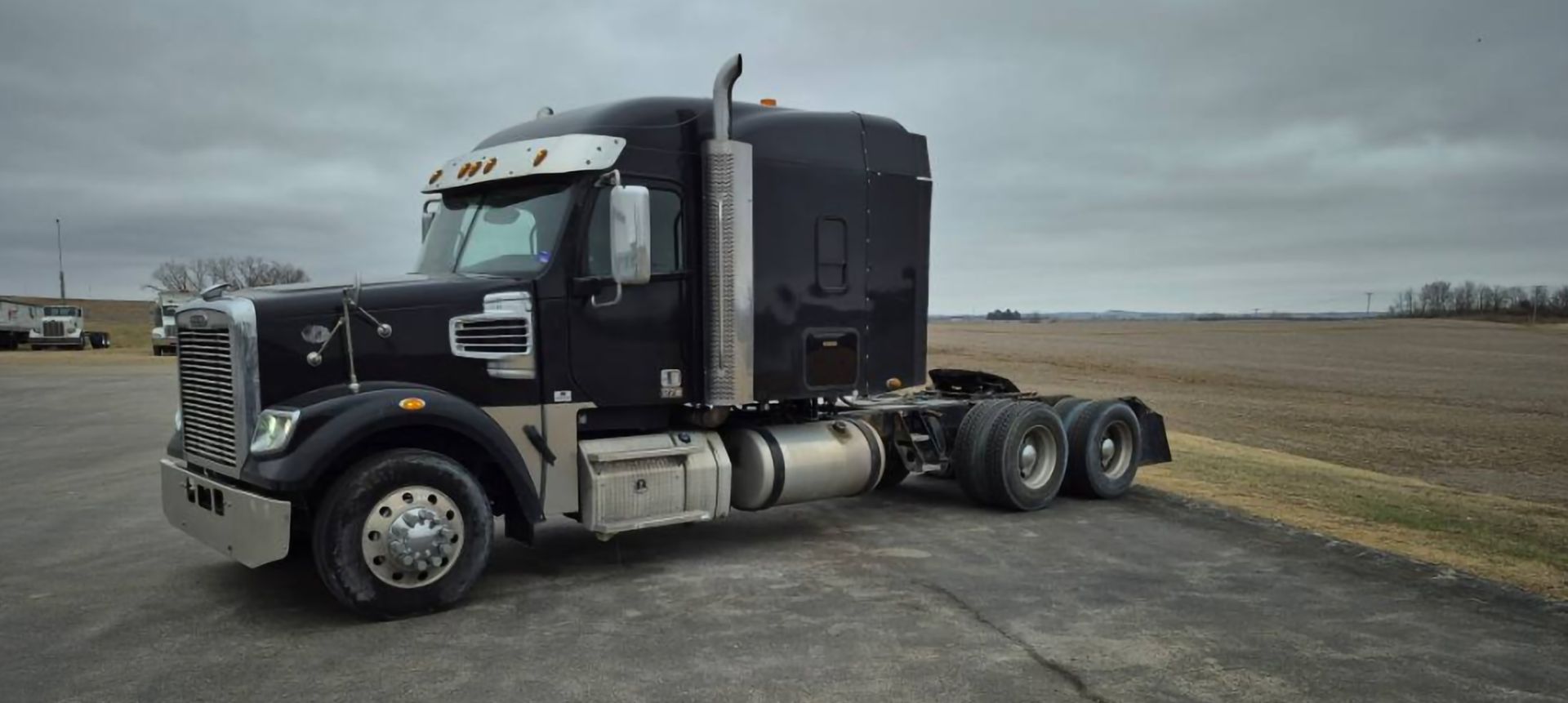 Black semi-truck on a gray day, parked on asphalt next to a field.
