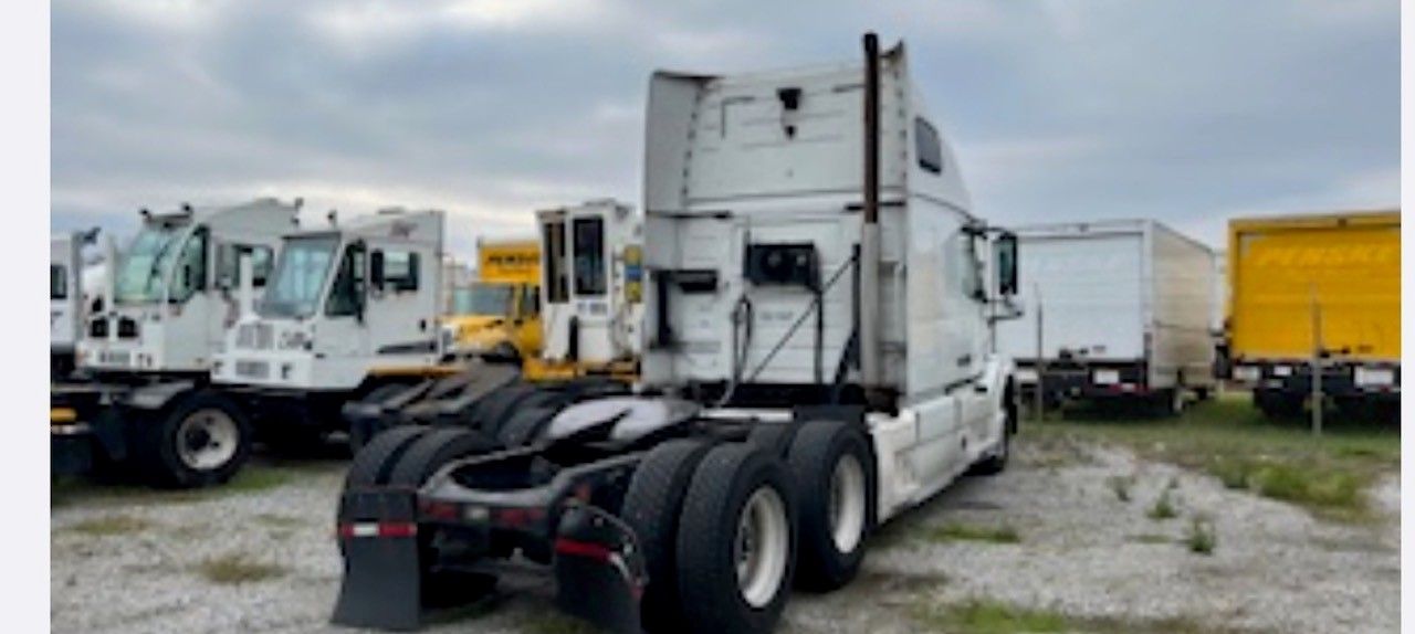 A white semi-truck parked outdoors. Other trucks and trailers are nearby. Cloudy sky in the background.