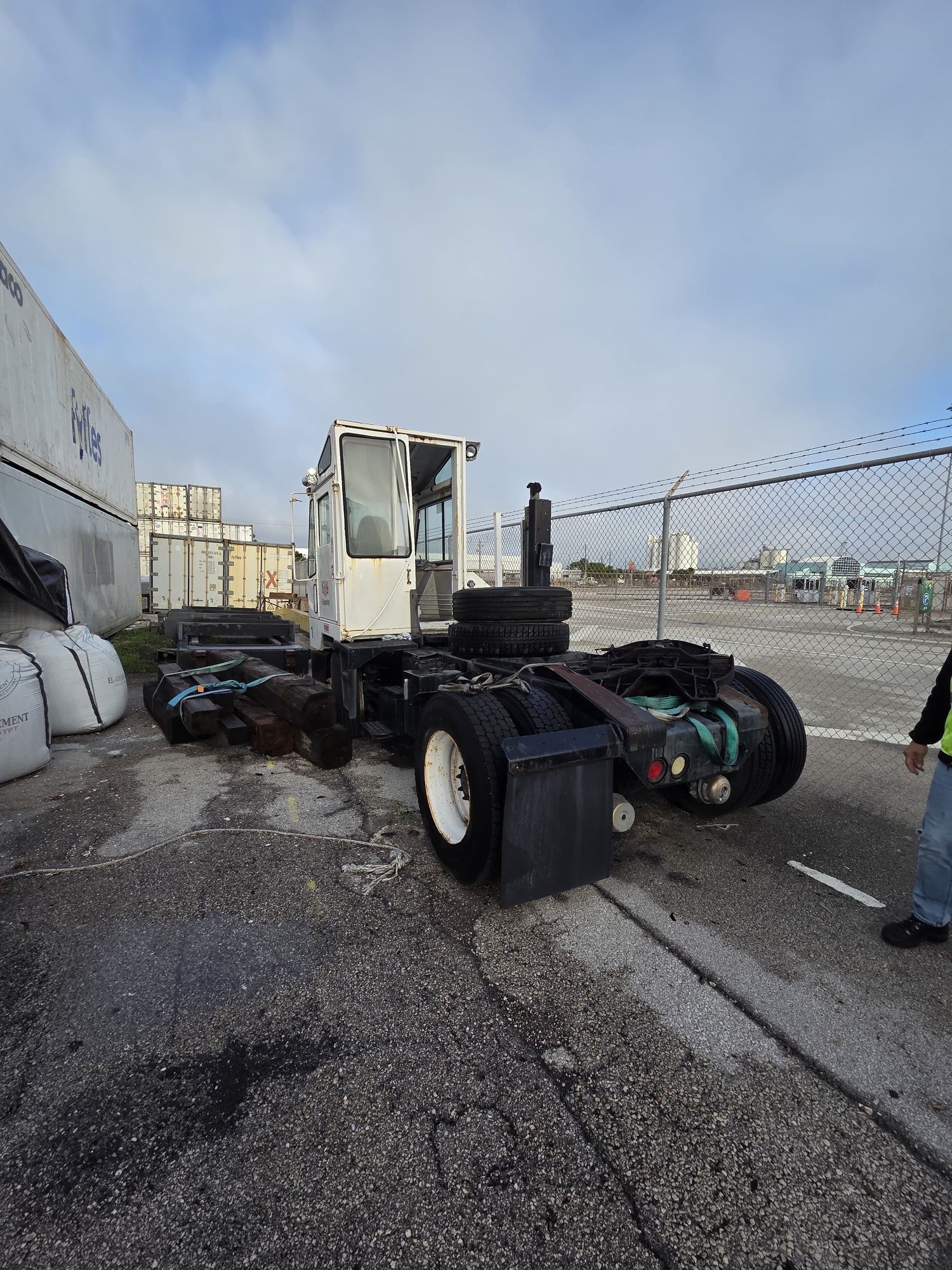 White yard truck on asphalt; next to cargo containers, fence. Cloudy sky in the background.