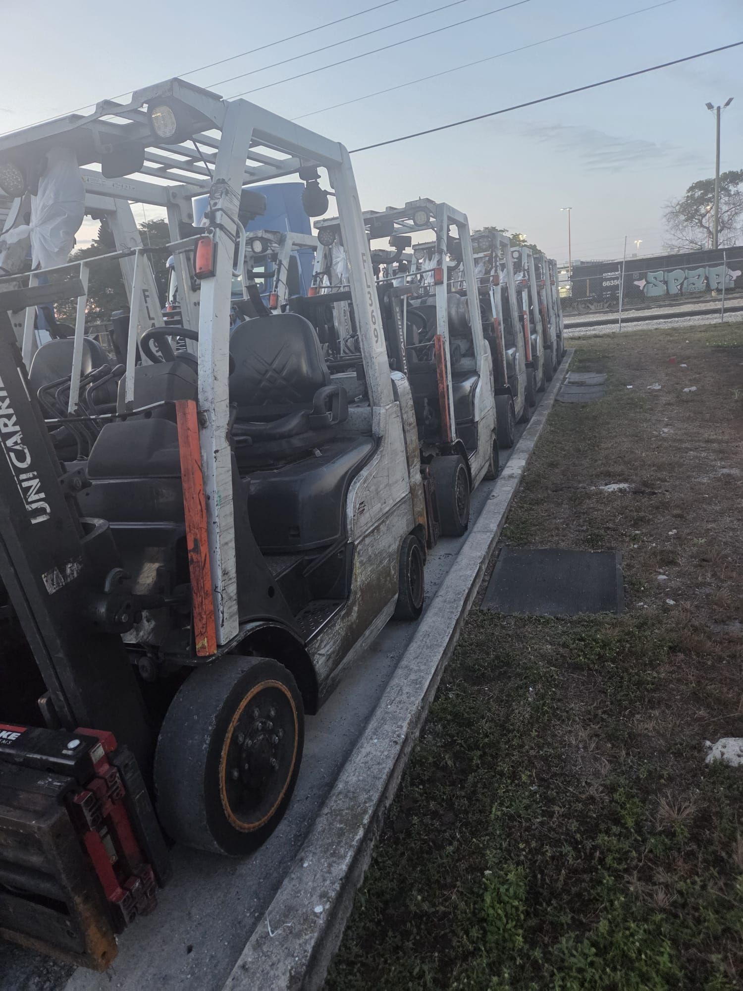 Row of white forklifts parked along a concrete barrier, under a cloudy sky.