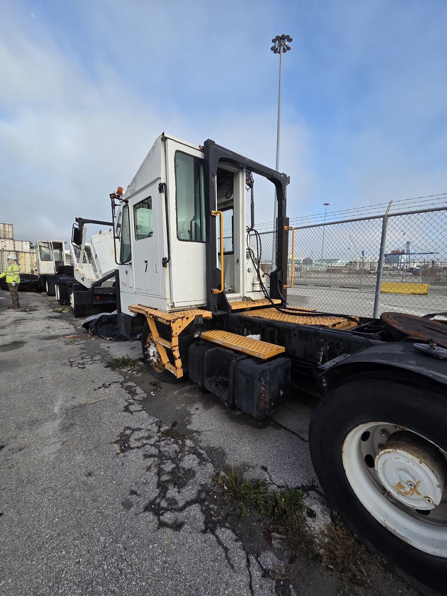 White yard truck with yellow accents parked on a paved lot next to a chain-link fence under a cloudy sky.