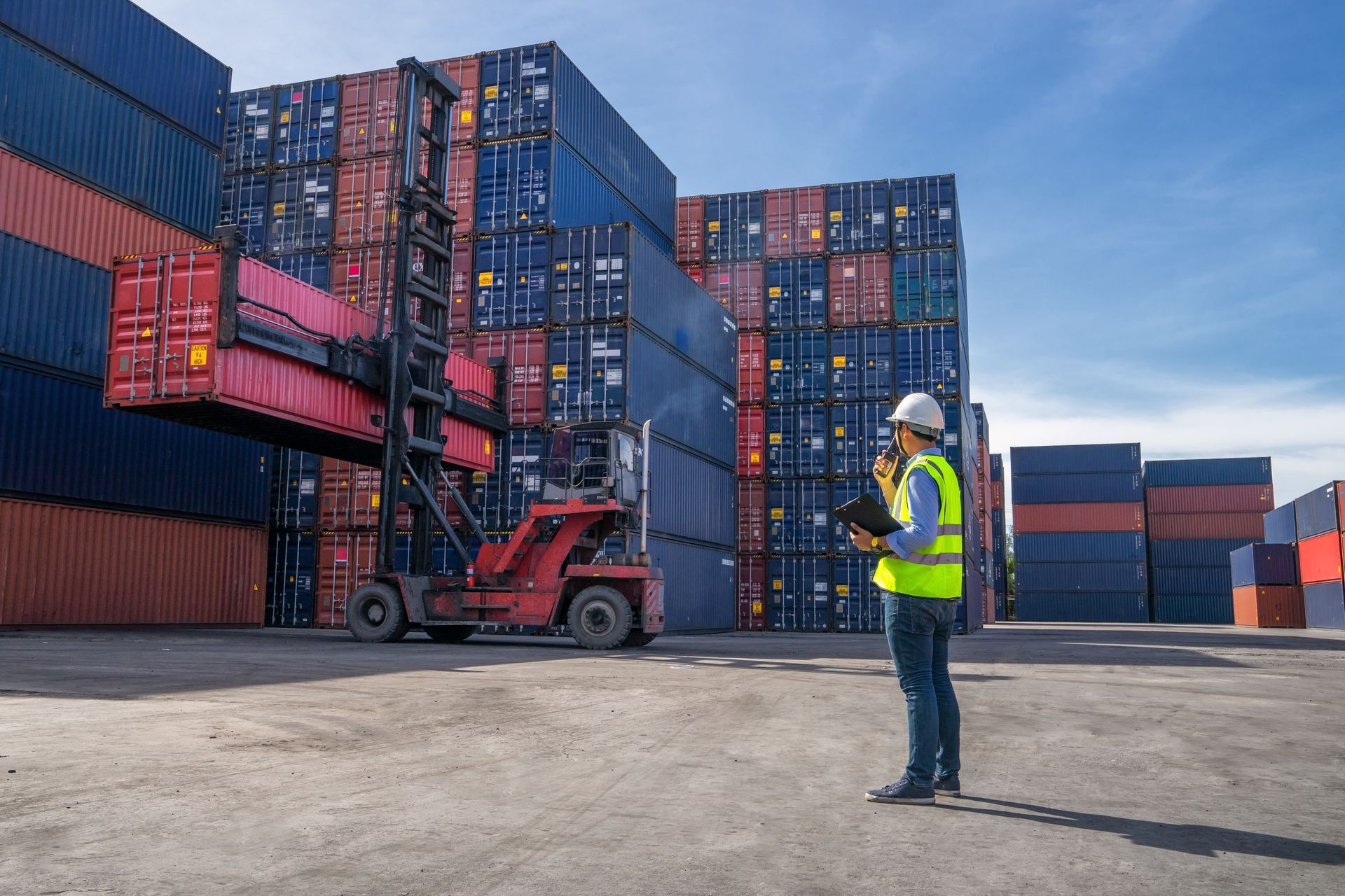 Forklift loading red shipping container in a port, overseen by worker in safety gear.