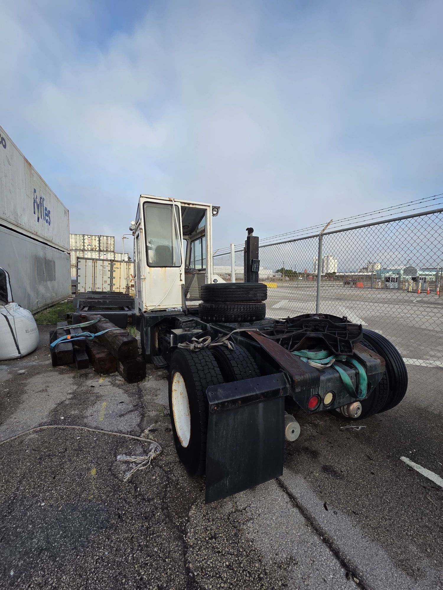 White semi-truck cab without trailer. Stacked tires and chain-link fence in industrial setting.