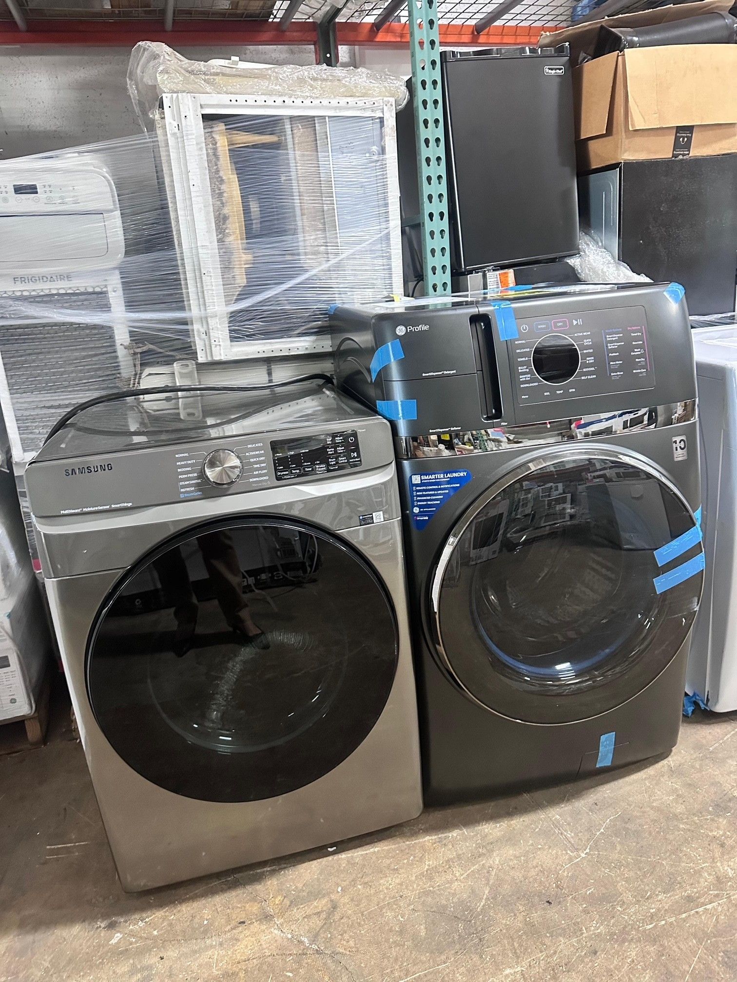 Two washing machines, one silver and one black, in a warehouse setting.