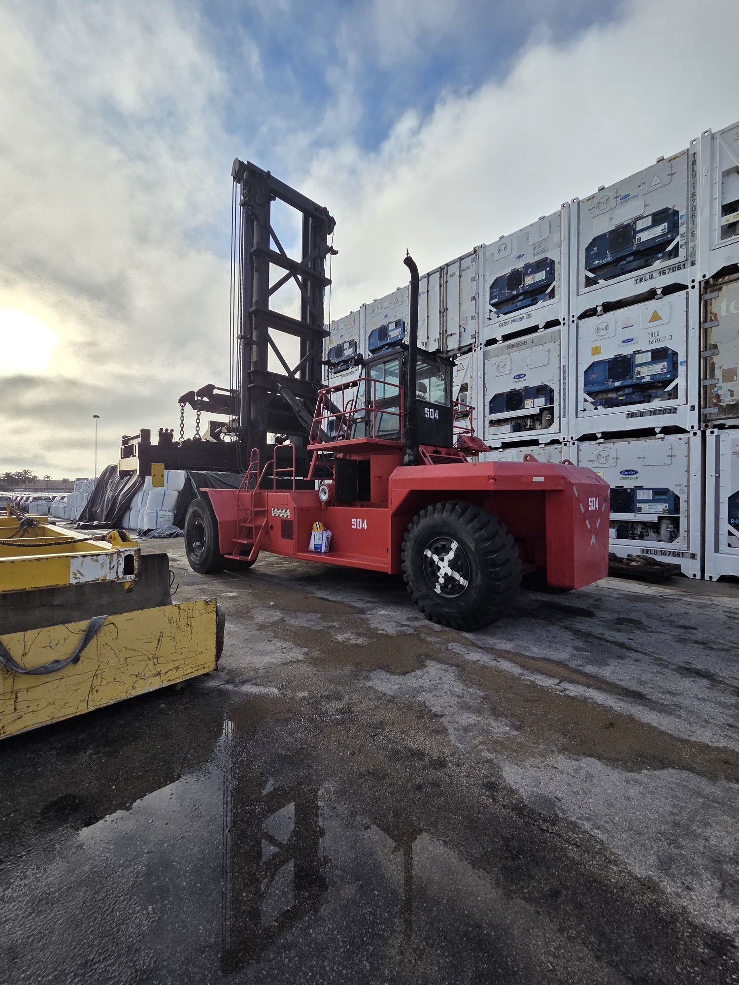 Red container handler at a port, lifting mechanism extended, beside stacked white shipping containers.