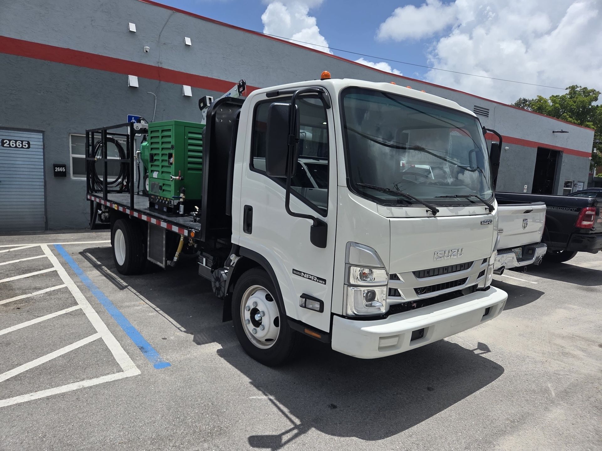 White Isuzu flatbed truck parked in front of a building with a green generator on the bed.