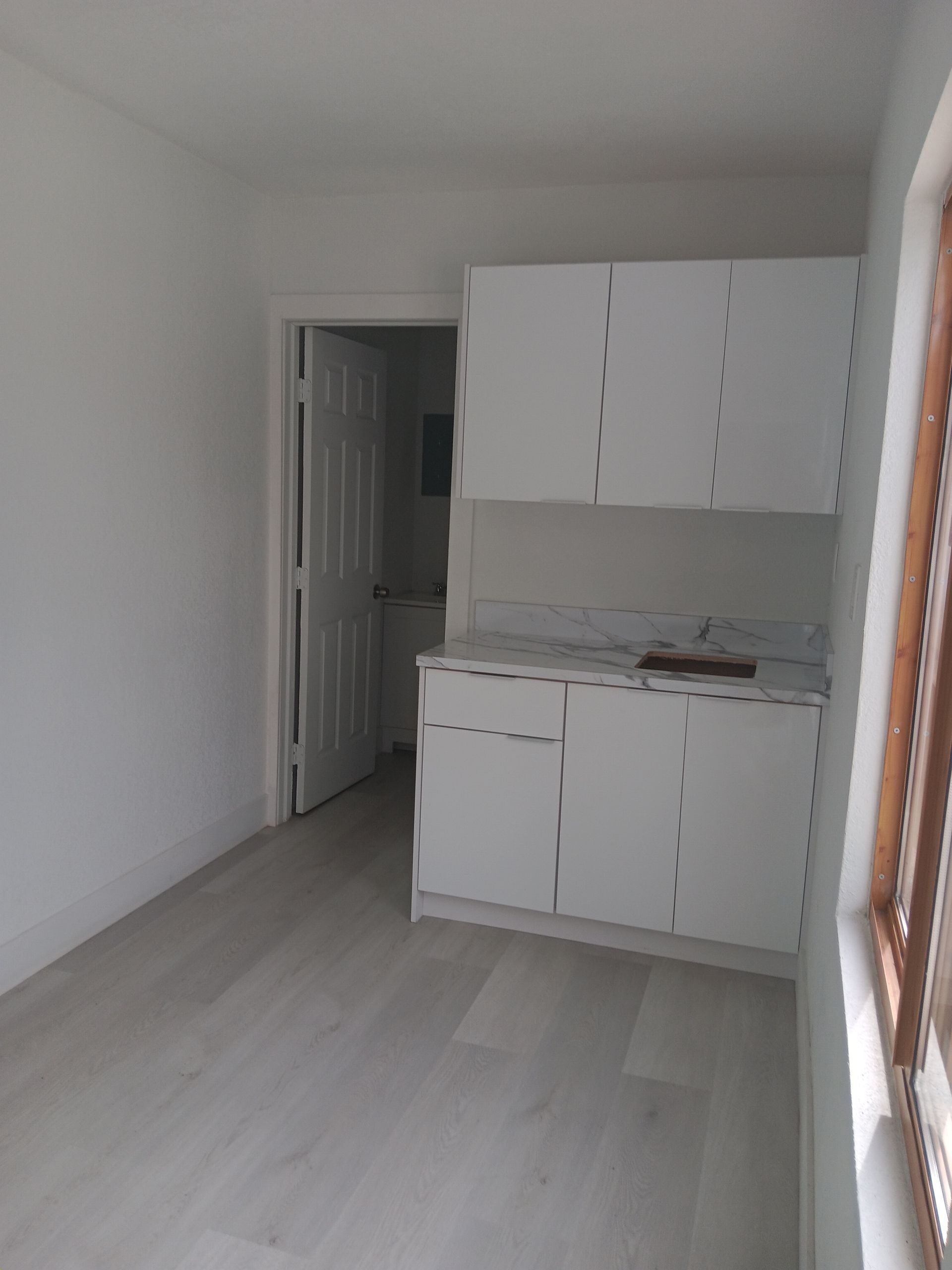 Small, white kitchenette with cabinets. Doorway to a bathroom is visible. Light-colored flooring and window.