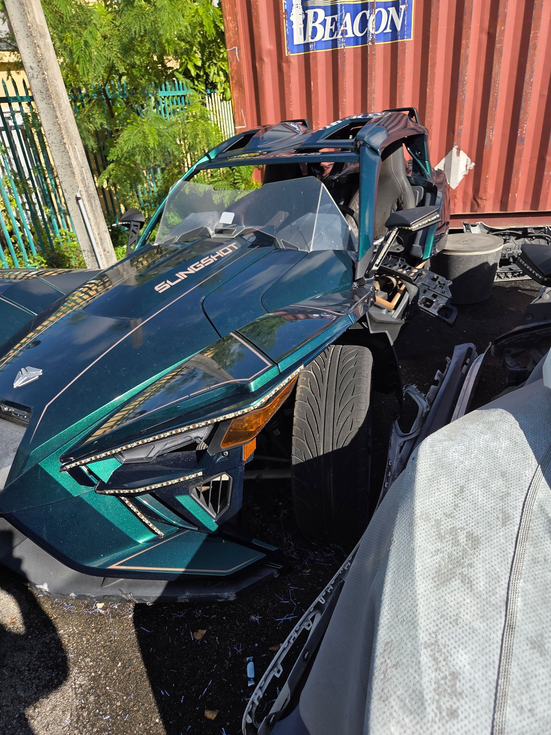 Green Polaris Slingshot vehicle with front-end damage; parked outdoors near a container.