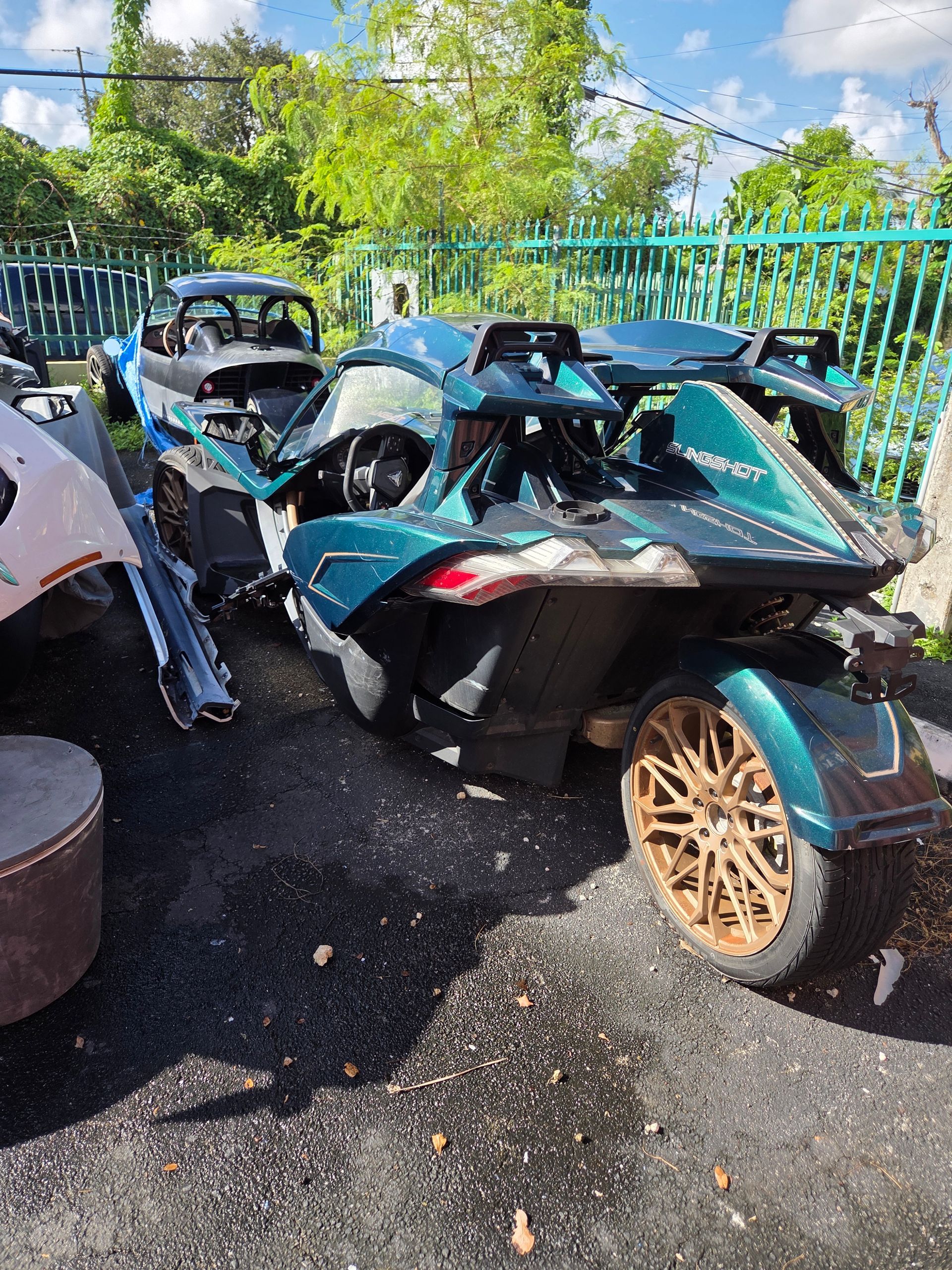 Damaged teal Polaris Slingshot three-wheeled vehicle, with visible body damage and gold wheels, outdoors.