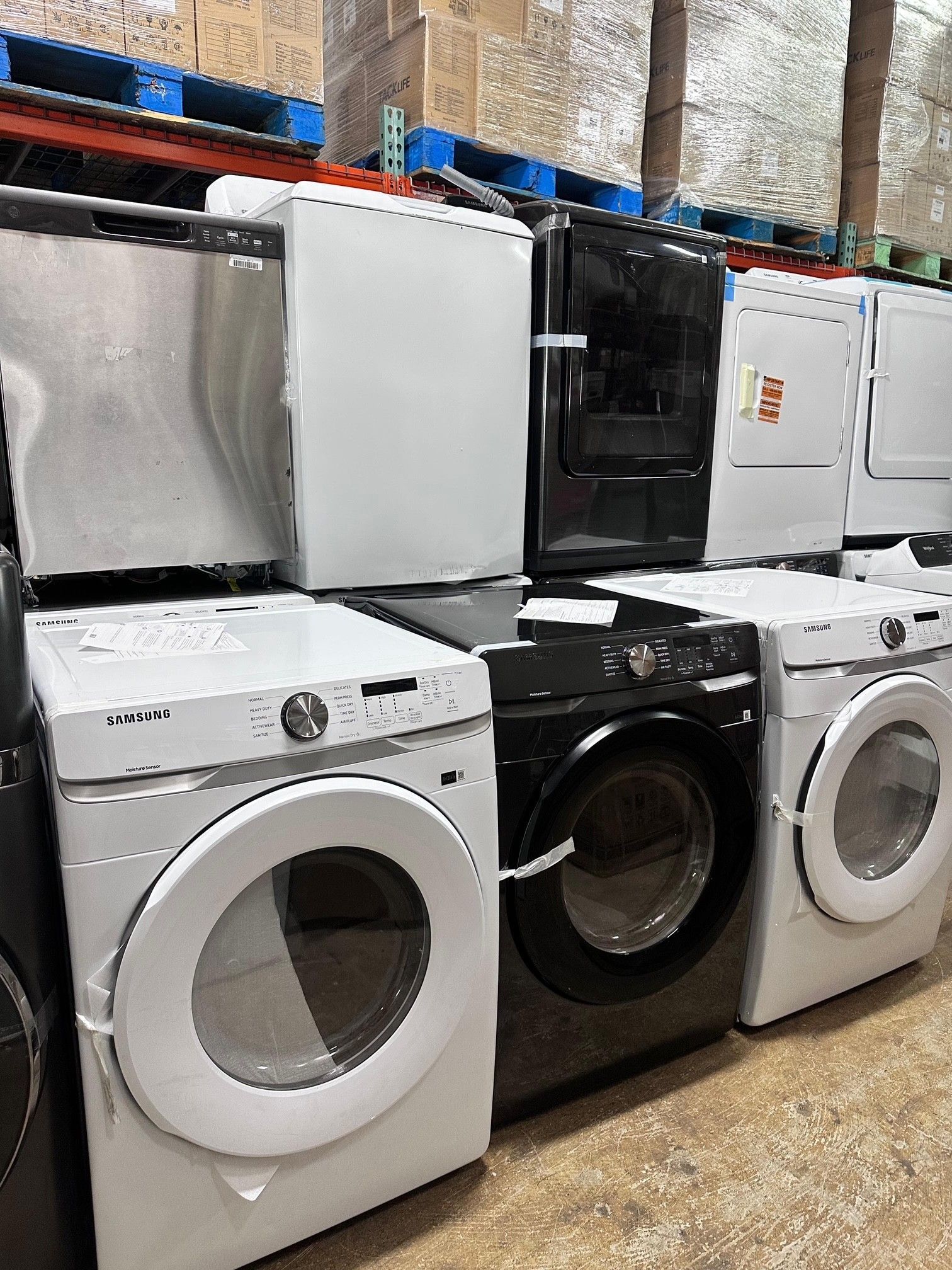Row of washers and dryers in various colors, including white, black, and stainless steel, in a warehouse setting.