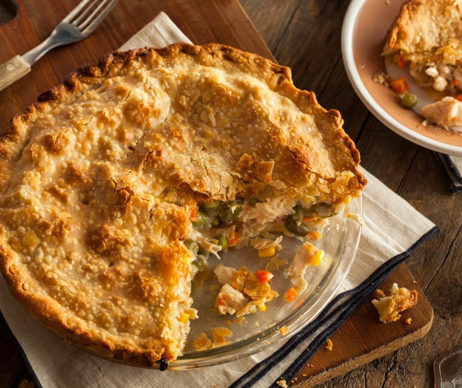Chicken pot pie in a glass pie dish, slice removed, on a wooden surface with silverware and a plate.