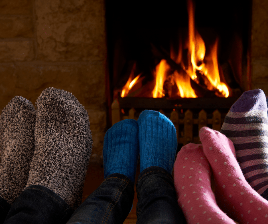 Four pairs of feet in colorful socks in front of a cozy fireplace with flames.