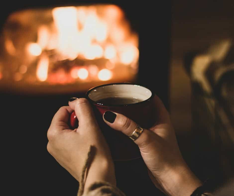 Hands holding a mug of tea in front of a warm fireplace.
