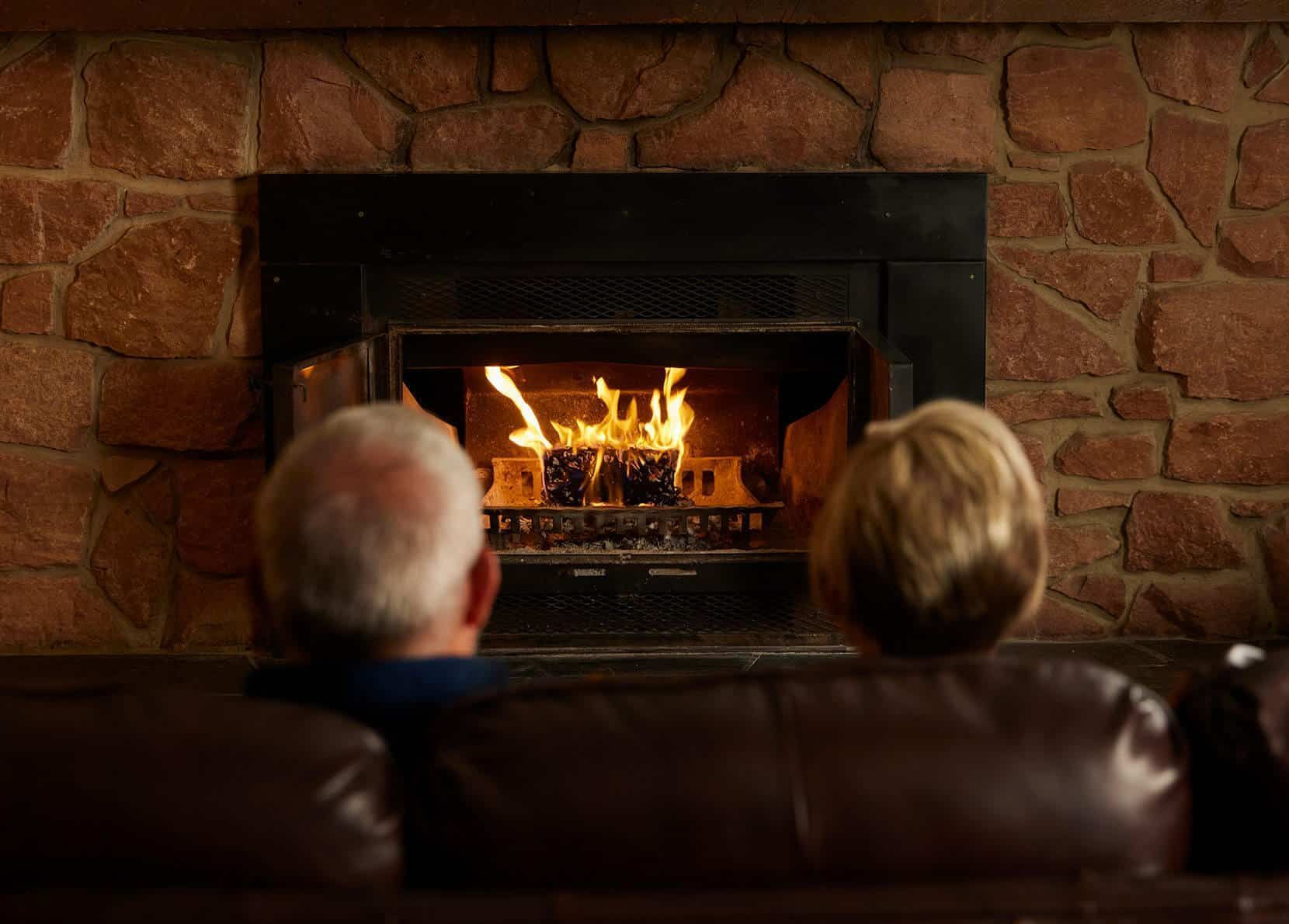 Two people sit on a brown sofa, facing a lit fireplace built into a stone wall.