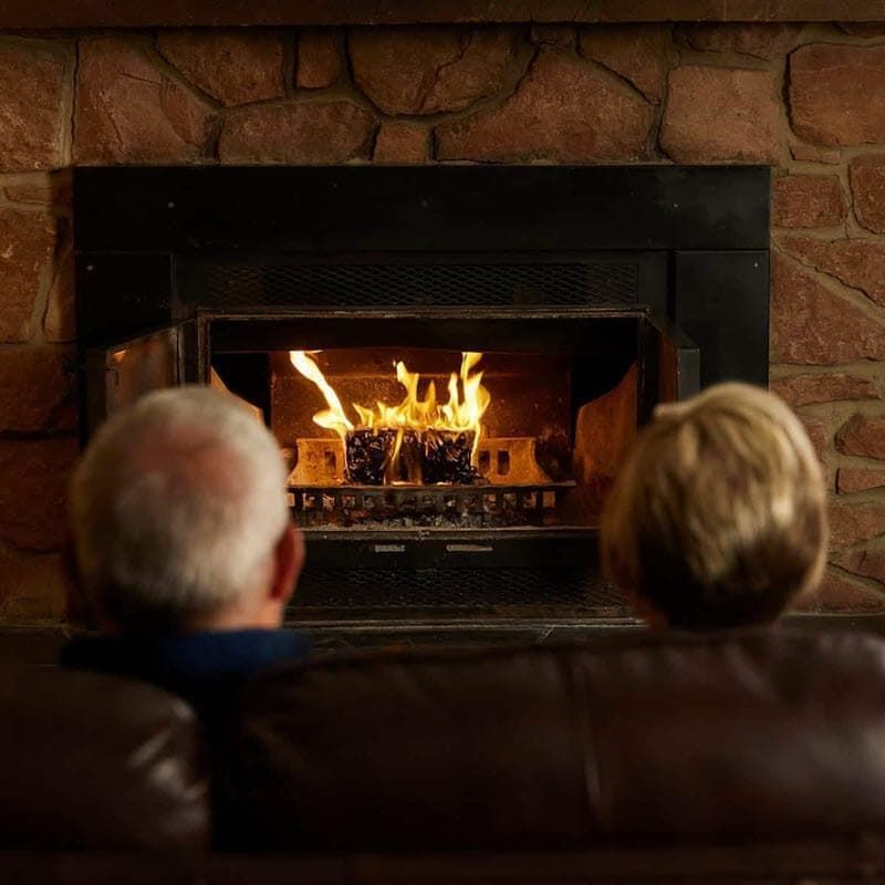 Two people sit on a couch, gazing at a fireplace with a burning fire in a stone-walled room.