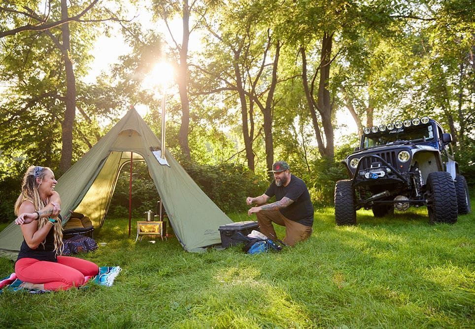 Couple camping near a modified Jeep. Person sits near tent, one prepares food outdoors on sunny day.