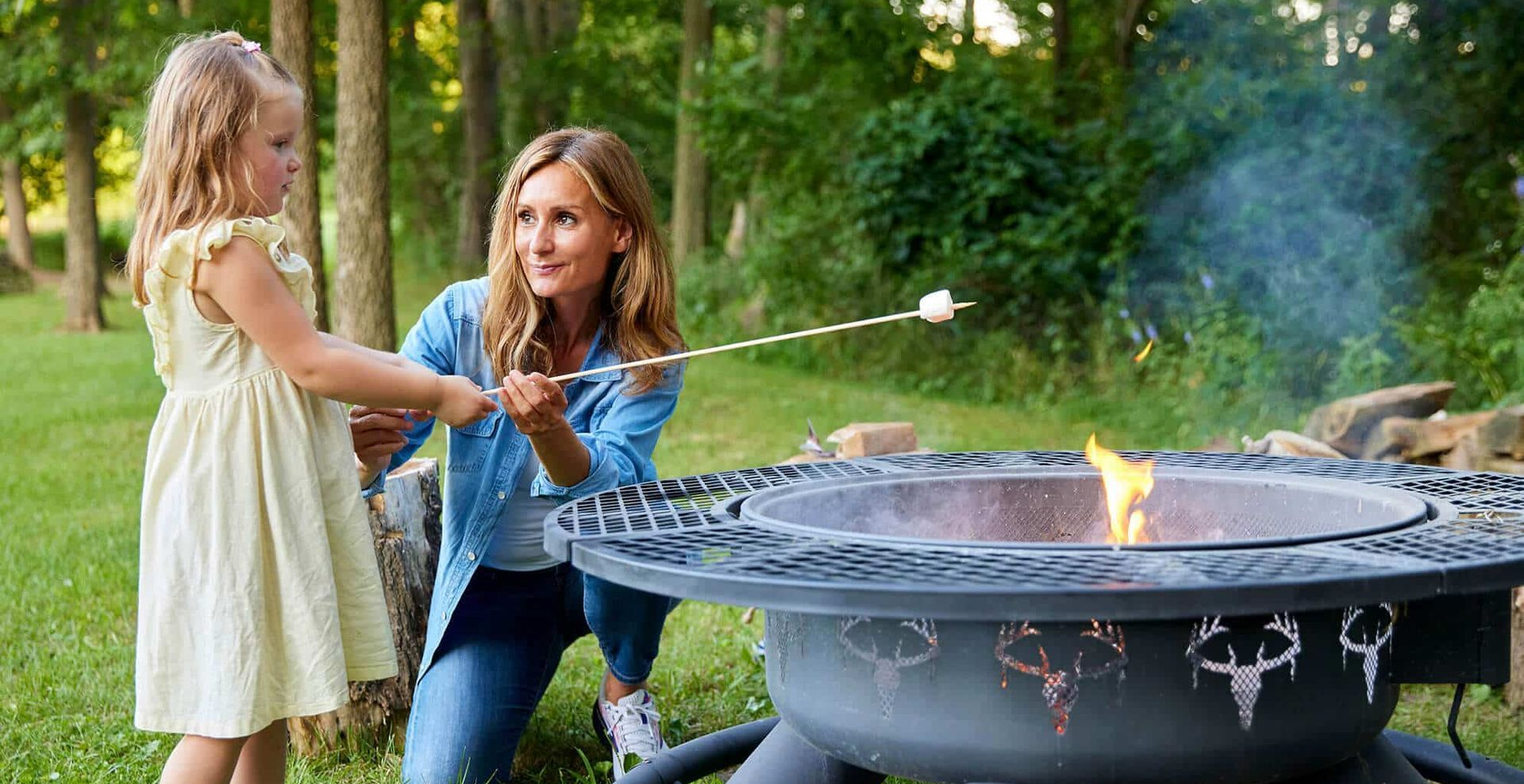 Woman and child roast a marshmallow over a fire pit in a grassy outdoor setting.