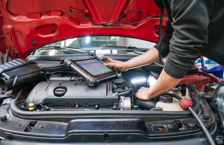 Person inspecting a car engine bay with a diagnostic tablet under the red hood