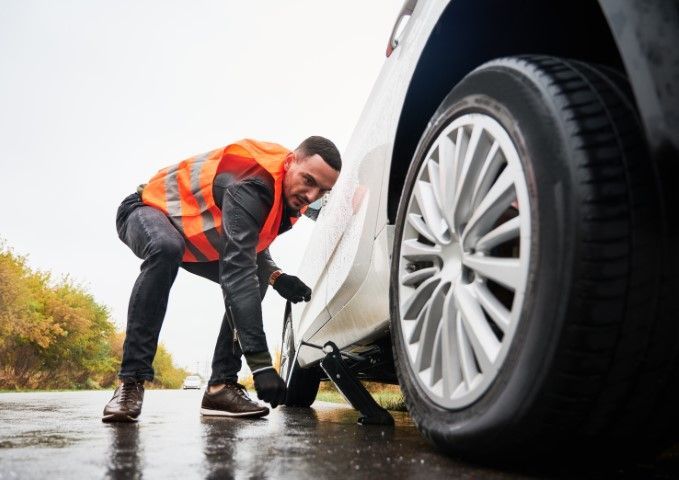 Person in an orange safety vest changing a car tire on a wet roadside