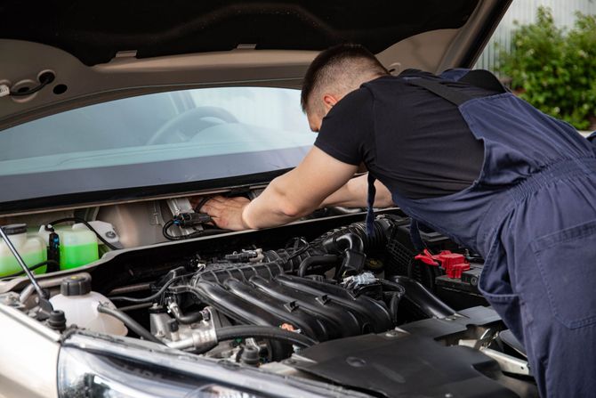 Mechanic in blue overalls working under a car hood in a garage