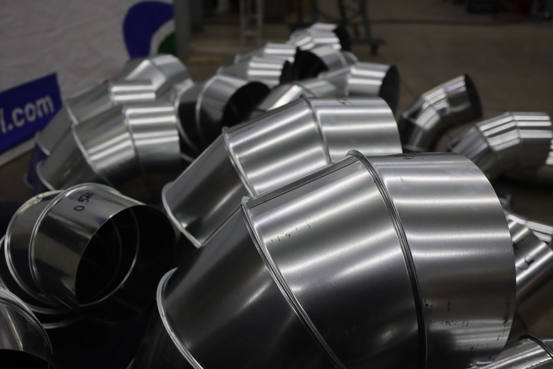 Silver metal ductwork elbows, stacked, in a factory setting.