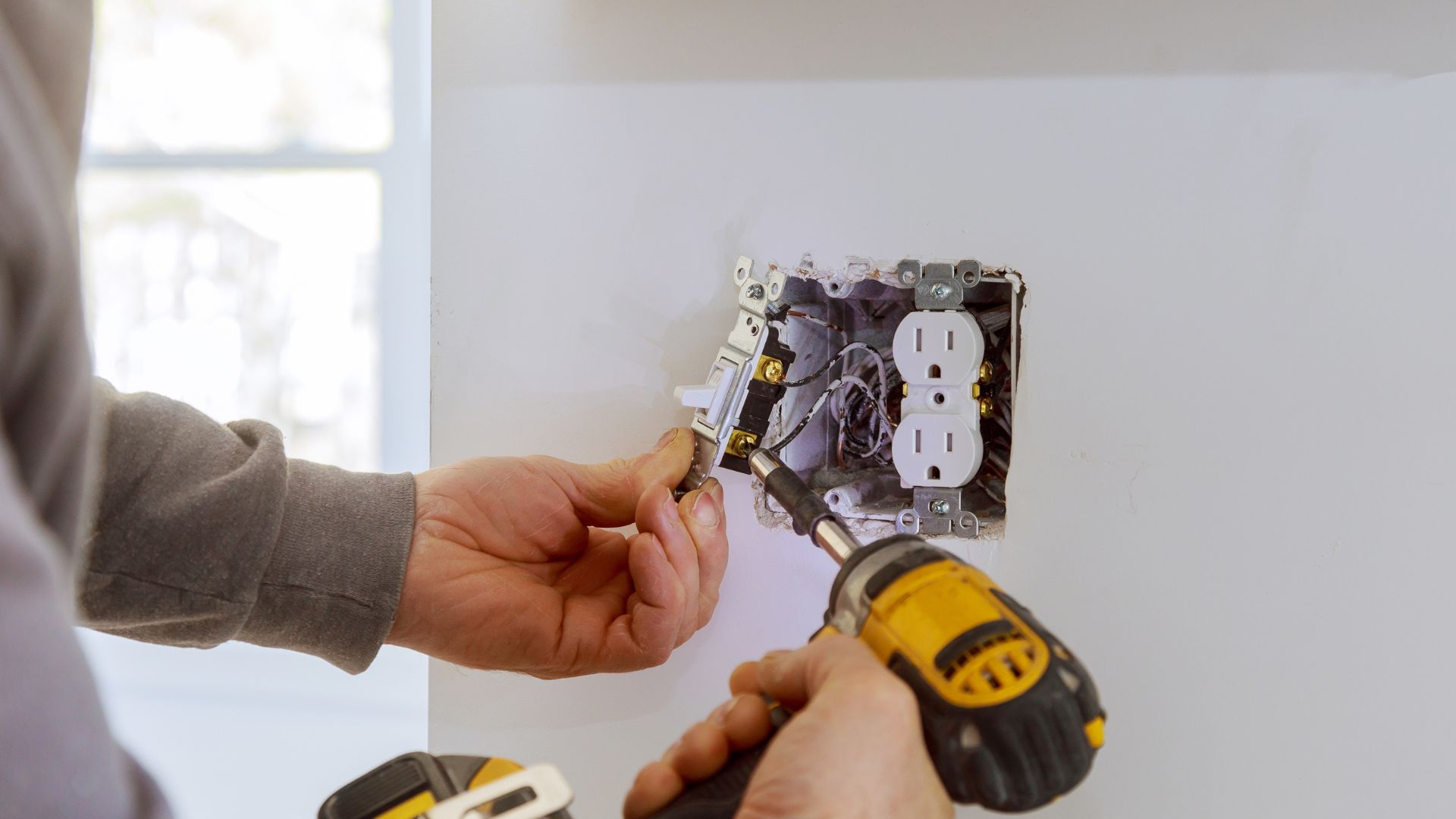 A person using a cordless power drill to secure a wall light switch and electrical outlet in a white room.