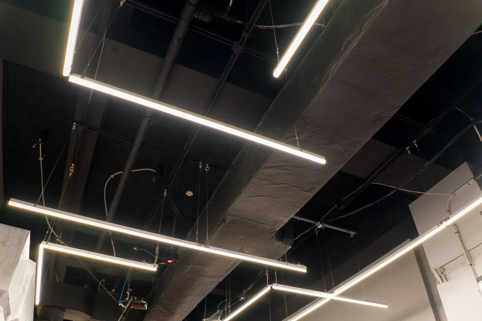 Linear LED light fixtures suspended from a dark, industrial-style ceiling with visible pipes and wiring.
