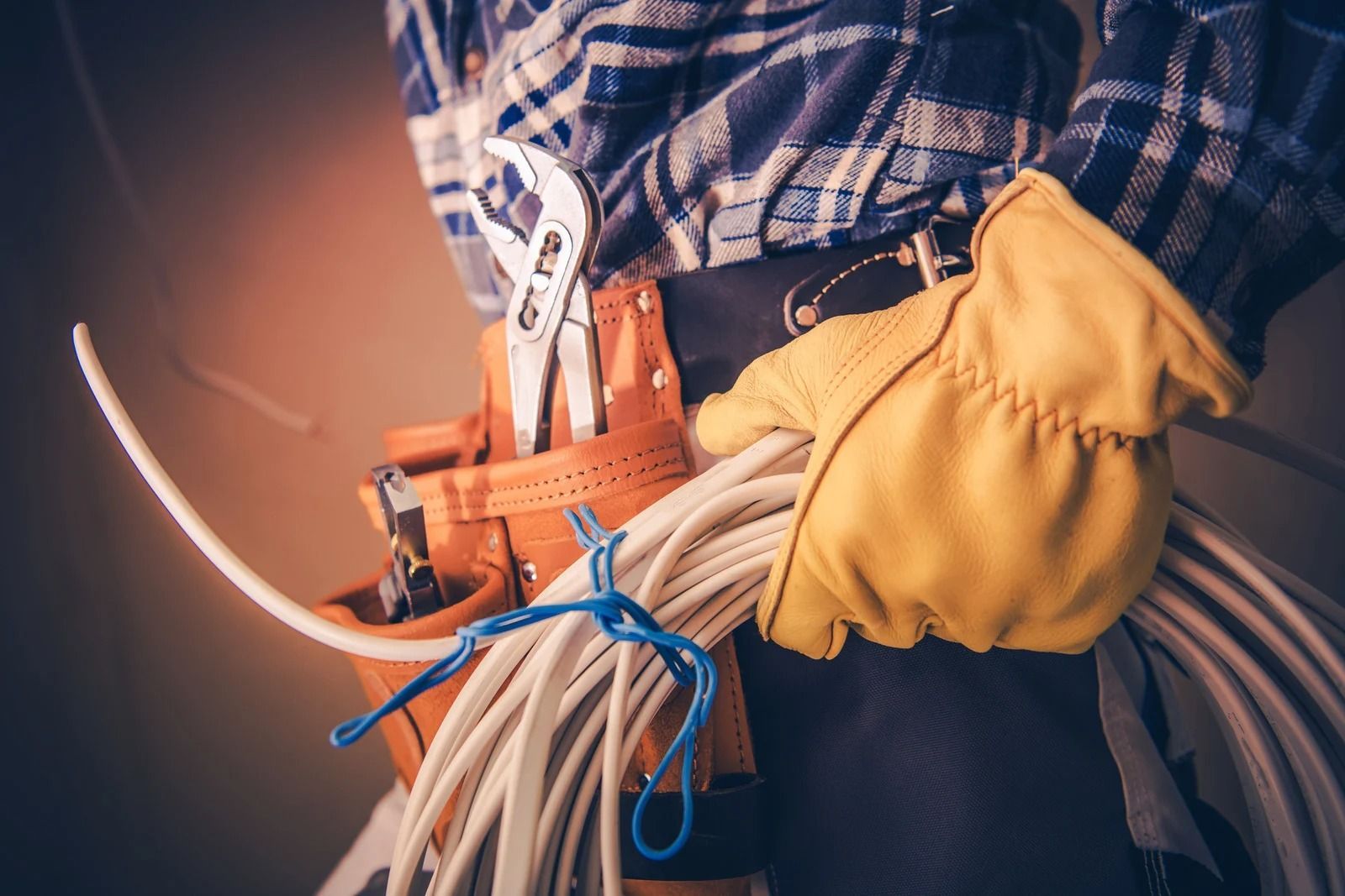 A worker in a plaid shirt and yellow work gloves carries a coil of electrical wire and a tool belt with pliers.