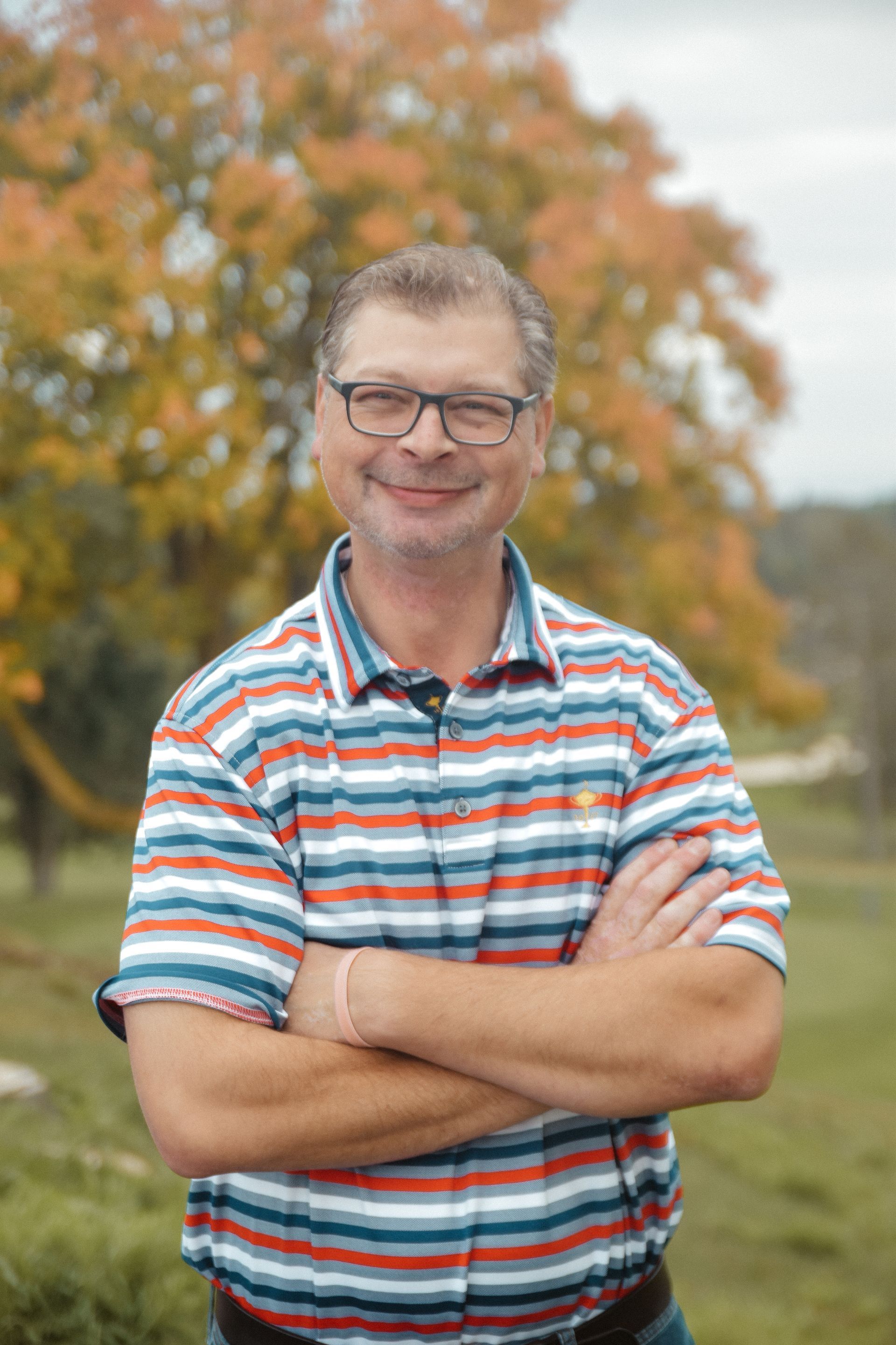 A man wearing glasses and a striped shirt is standing in a field with his arms crossed.