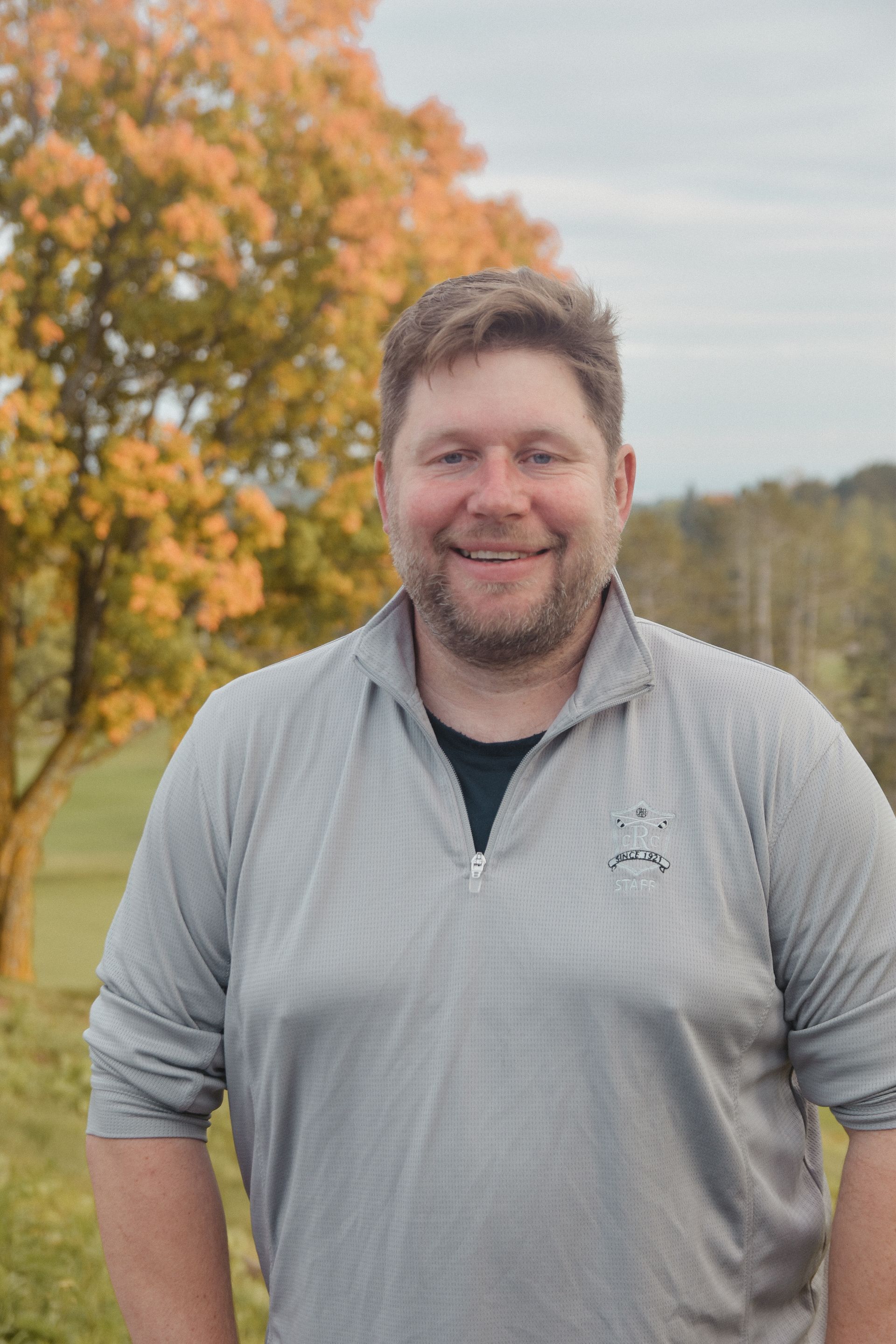A man with a beard is smiling for the camera while standing in front of a tree.