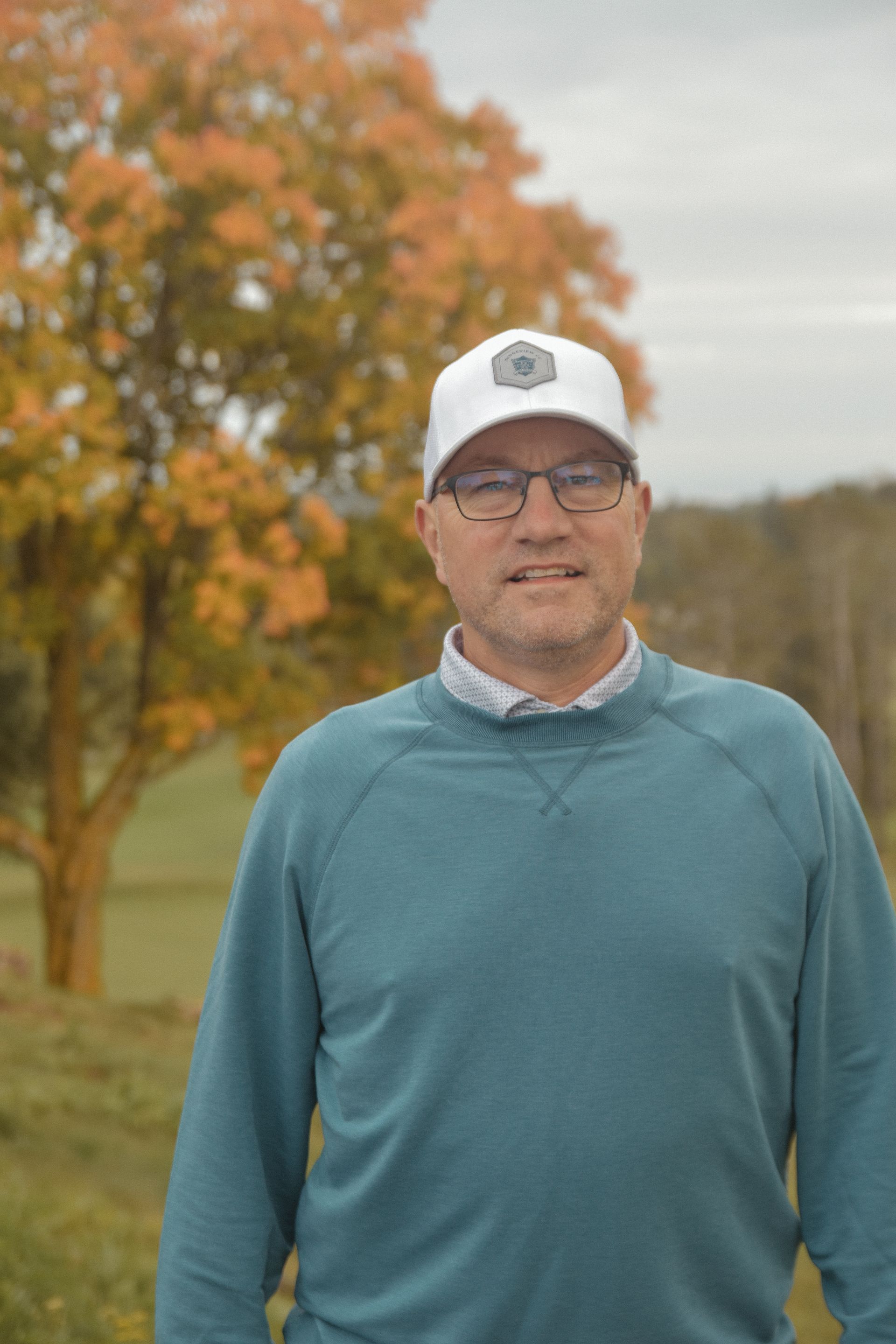 A man wearing a blue sweater and a white hat is standing in front of a tree.
