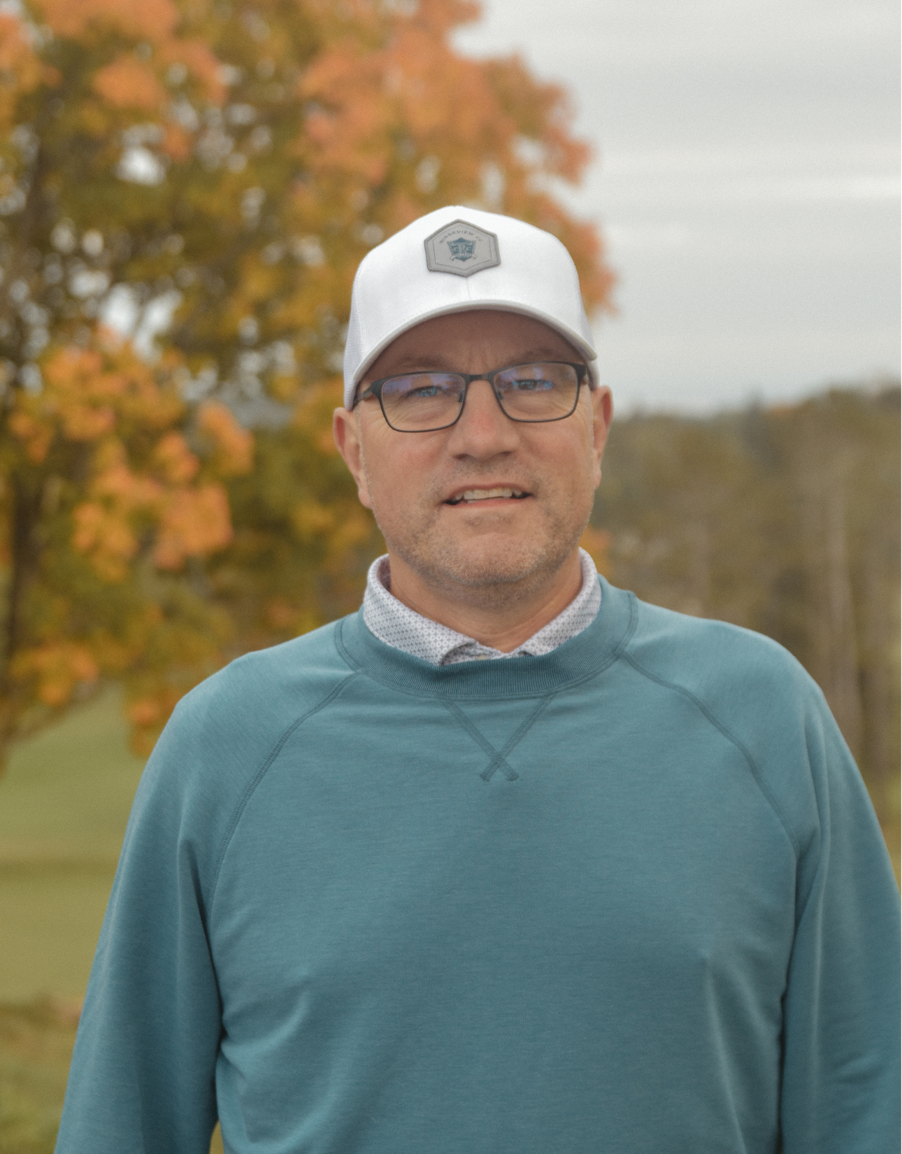 A man wearing a blue sweater and a white hat is standing in front of a tree.