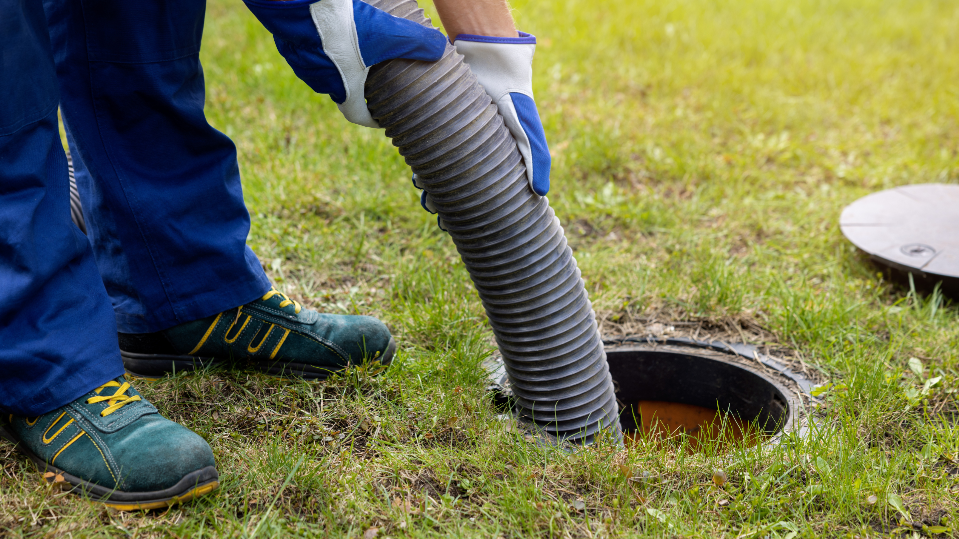 Person in blue overalls and gloves inserting a hose into a septic tank in a grassy yard.