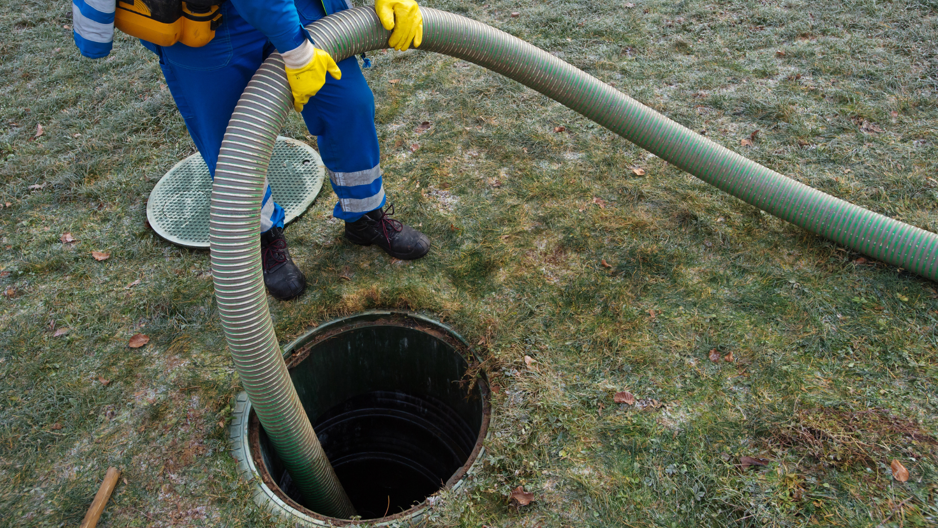 Person in blue coveralls and gloves empties a septic tank with a hose outdoors.