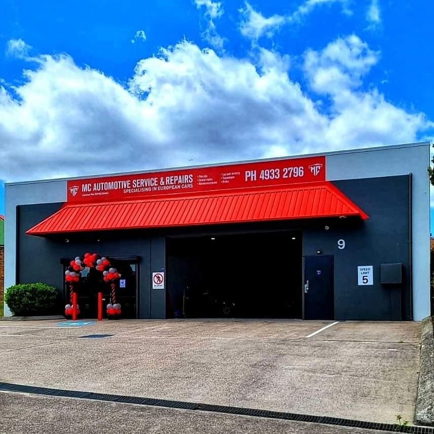 Red-roofed auto repair shop with an open garage door under a partly cloudy sky; balloons on the left.
