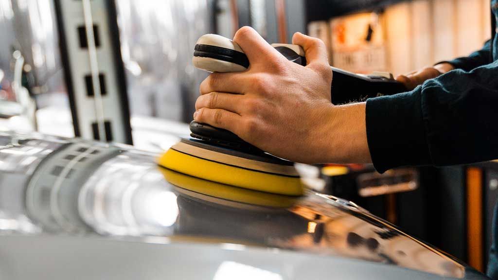 Person polishing a car with an electric buffer in a garage. Yellow and black buffing pad in use.