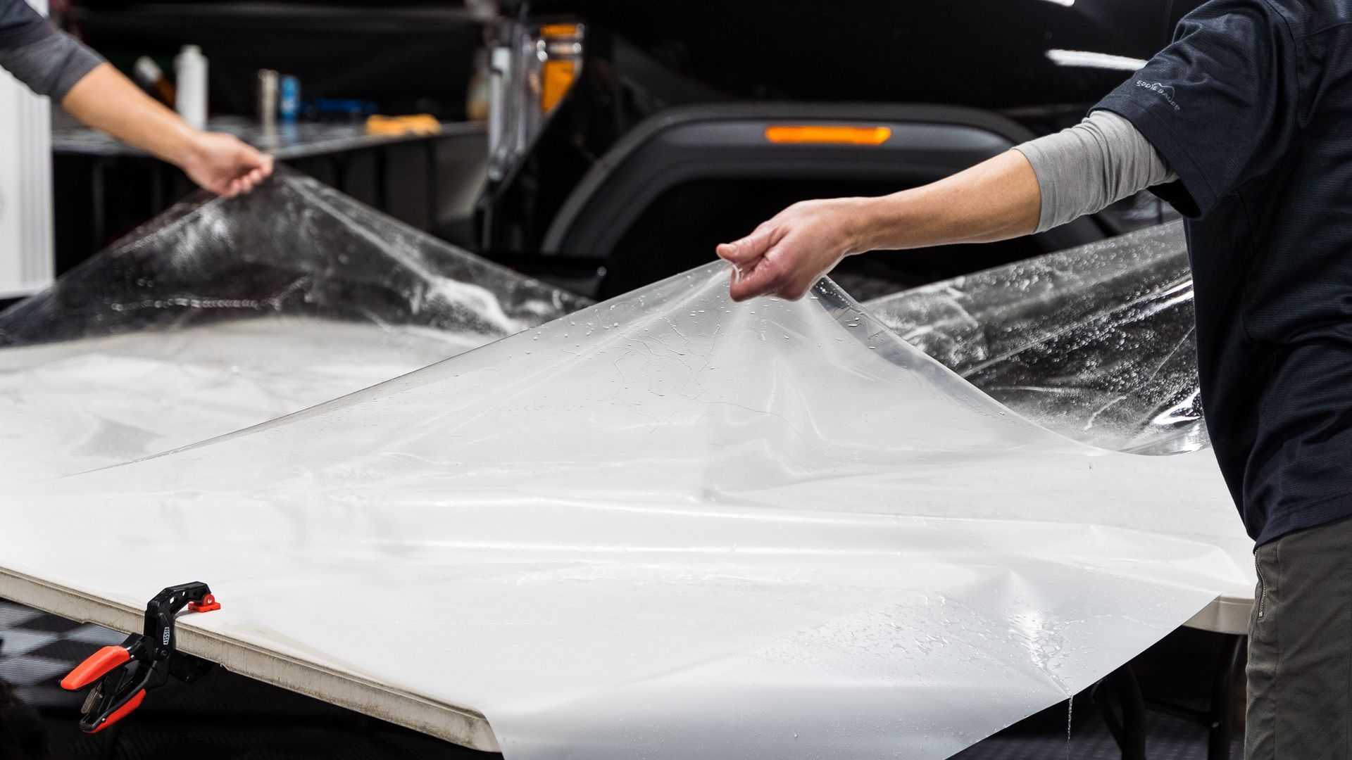 Hands holding protective film over a car hood. Indoors, shop setting.