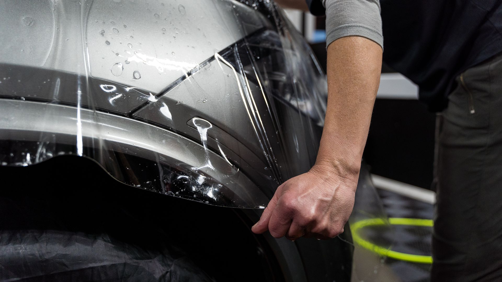 Person applying clear protective film to a car bumper. Water droplets visible.